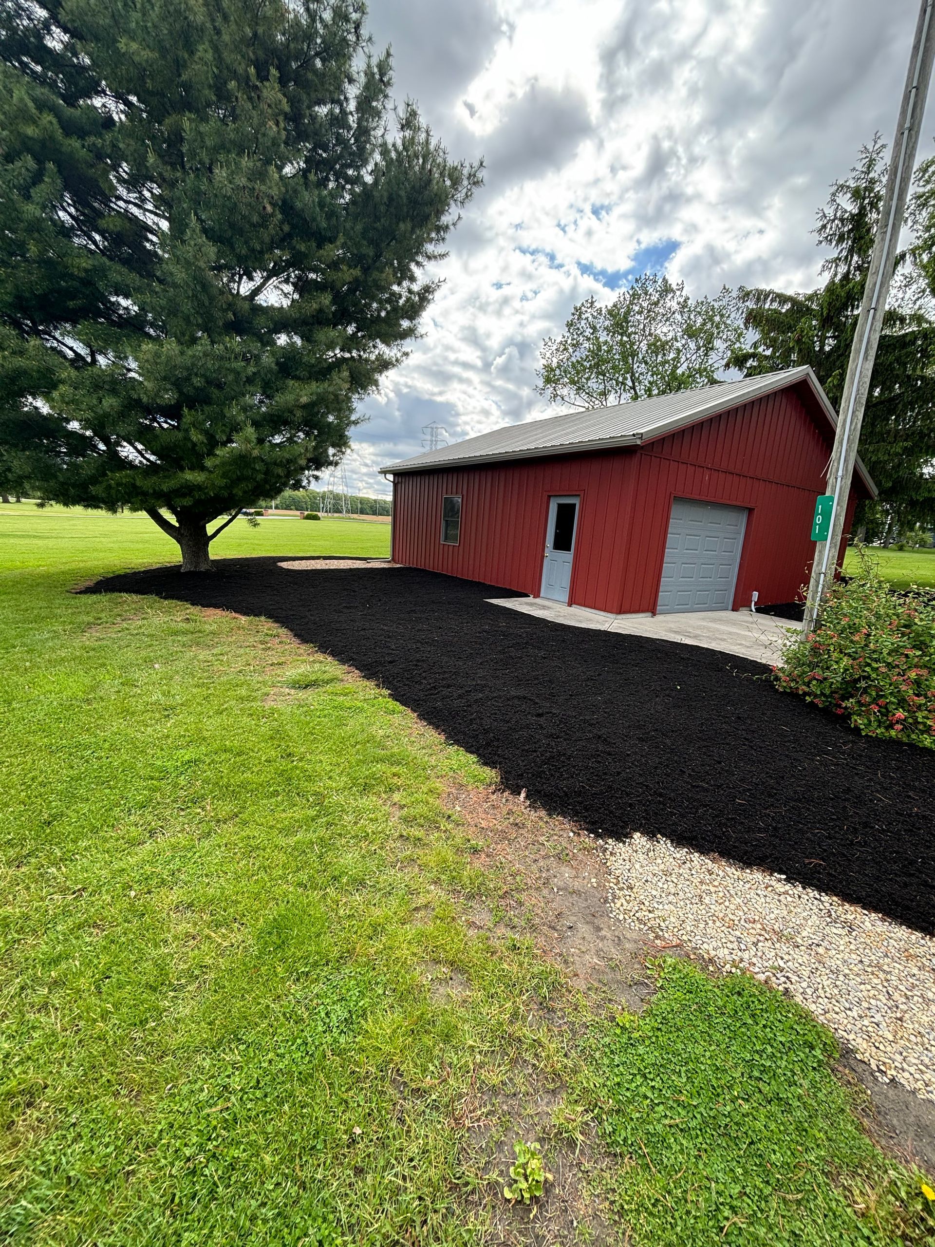 Black mulched path leads to a red shed with a gray door and garage, beside a large tree on green grass.