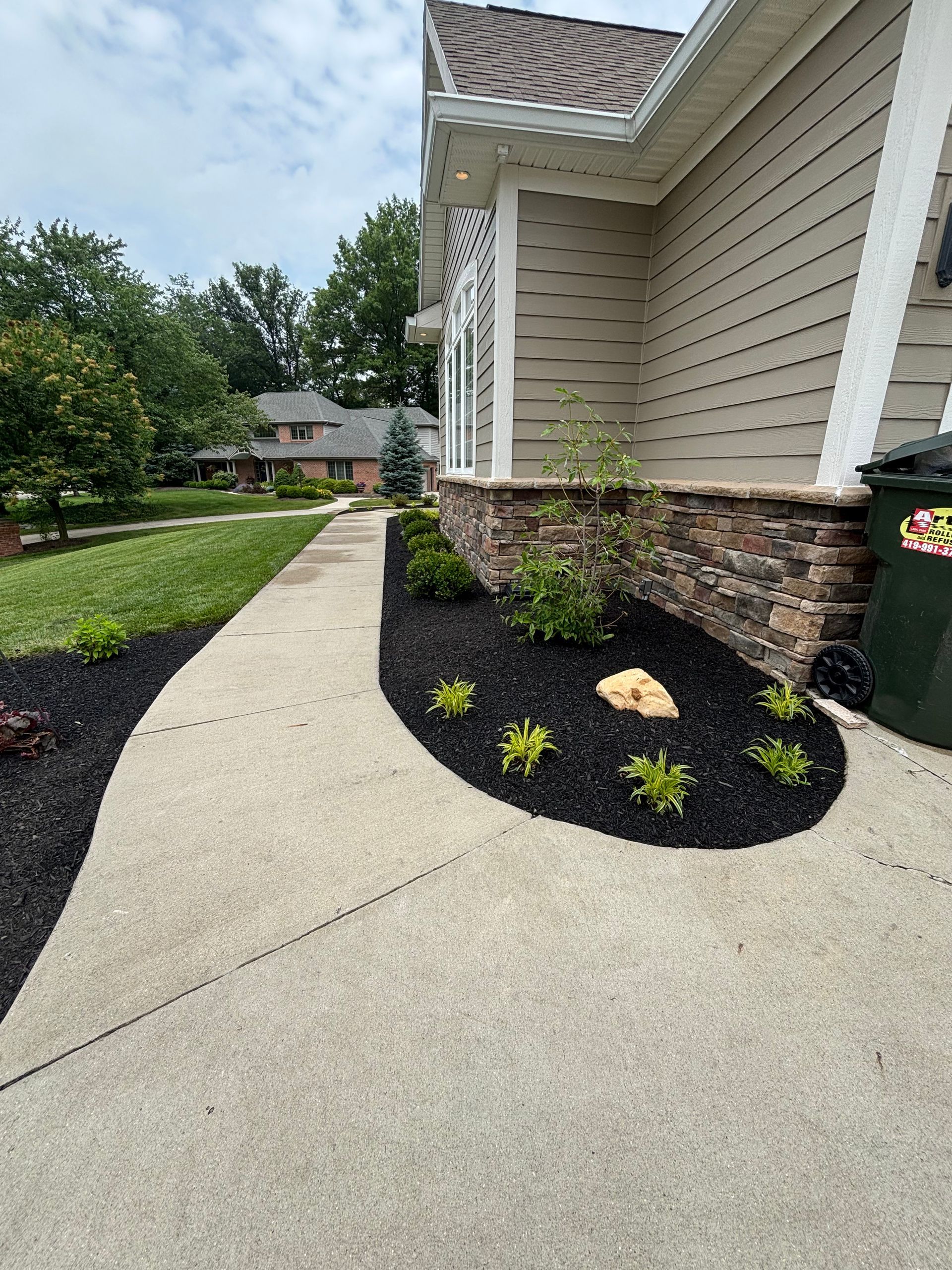 Concrete walkway curves past house with stone wall and flower bed with black mulch.