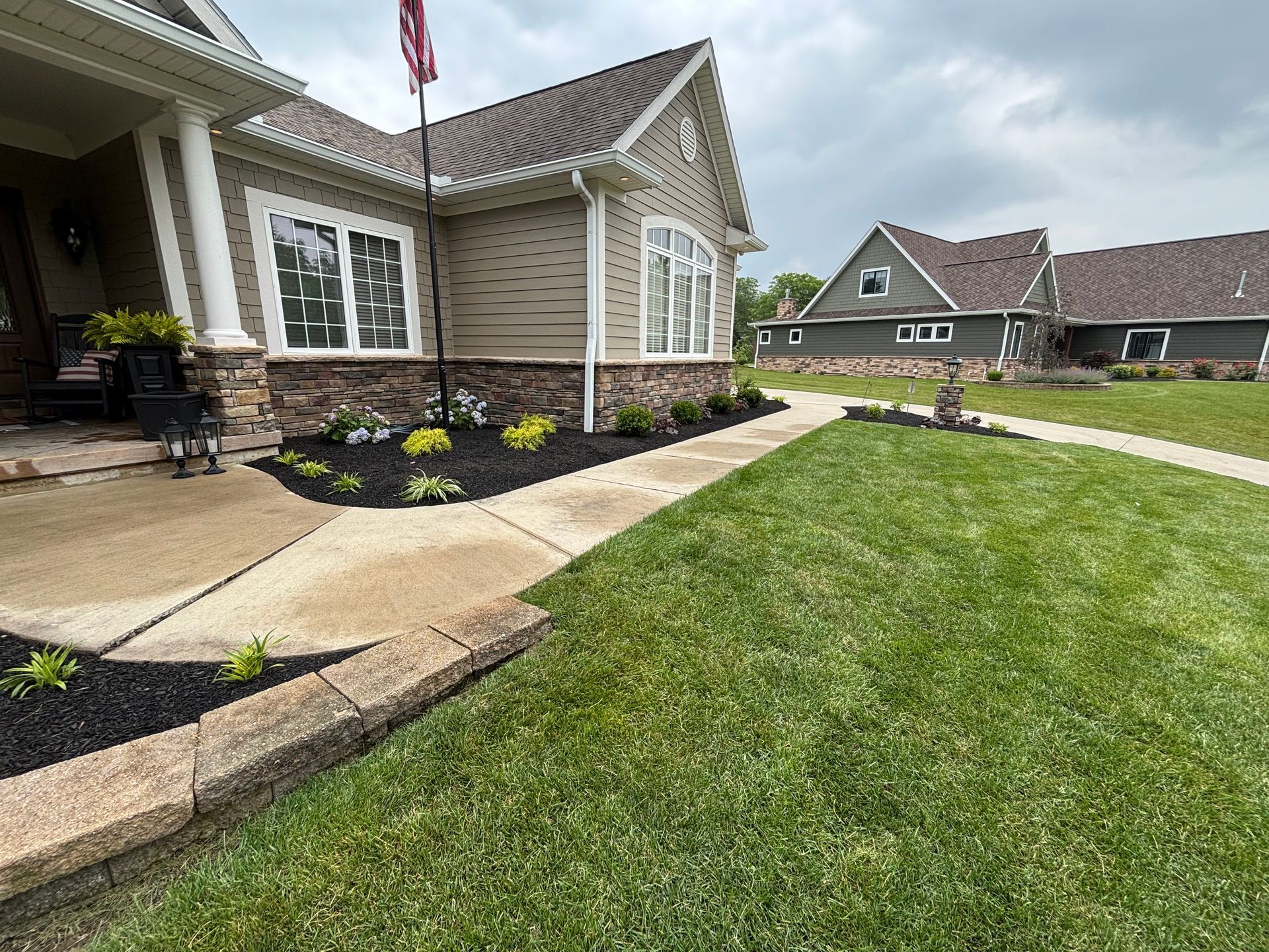 A house with a walkway and lawn, featuring fresh landscaping, flowers, and a flag on a cloudy day.