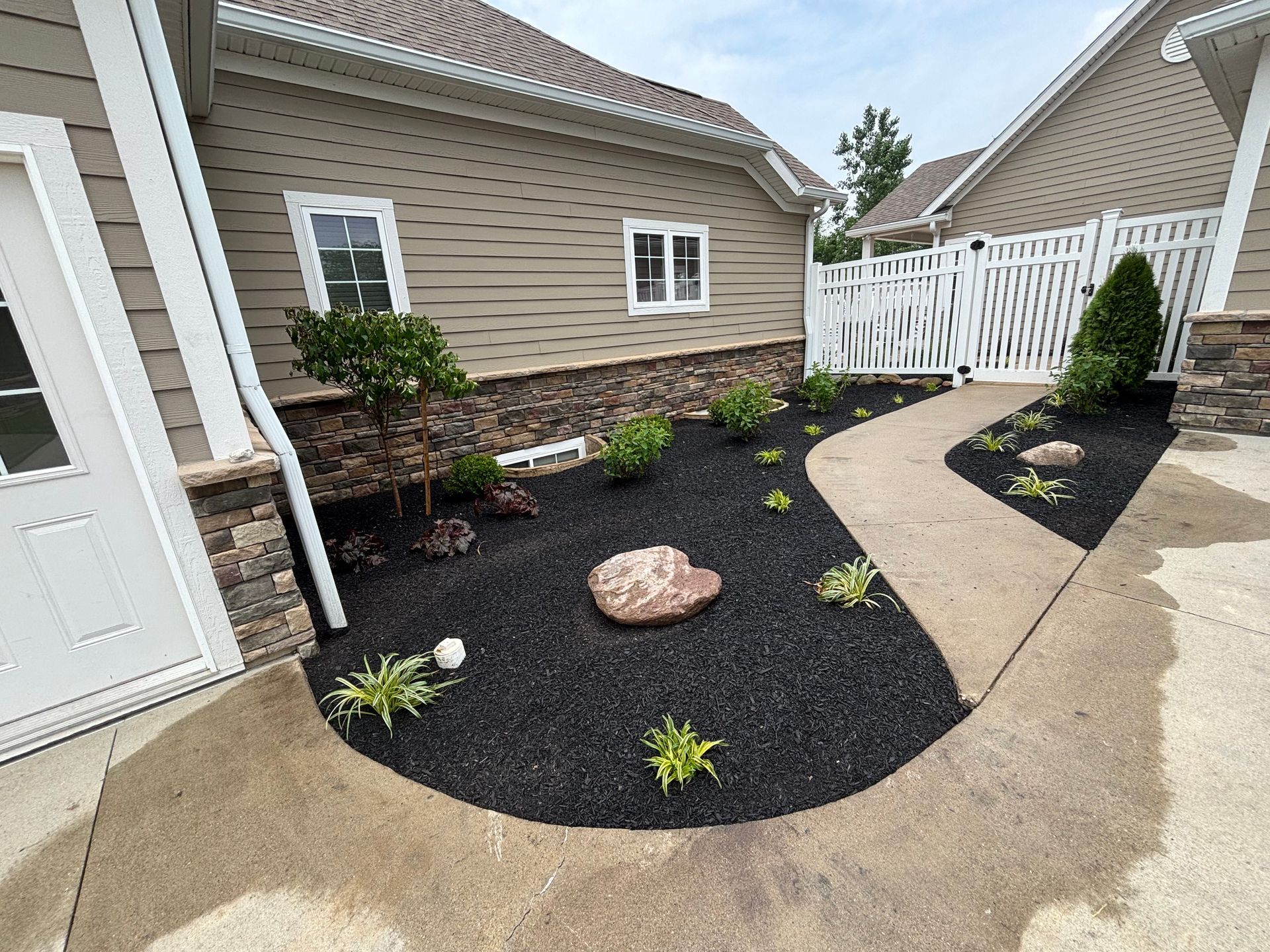 Landscaped bed with dark mulch, plants, and a stone, next to a tan house with a white fence.