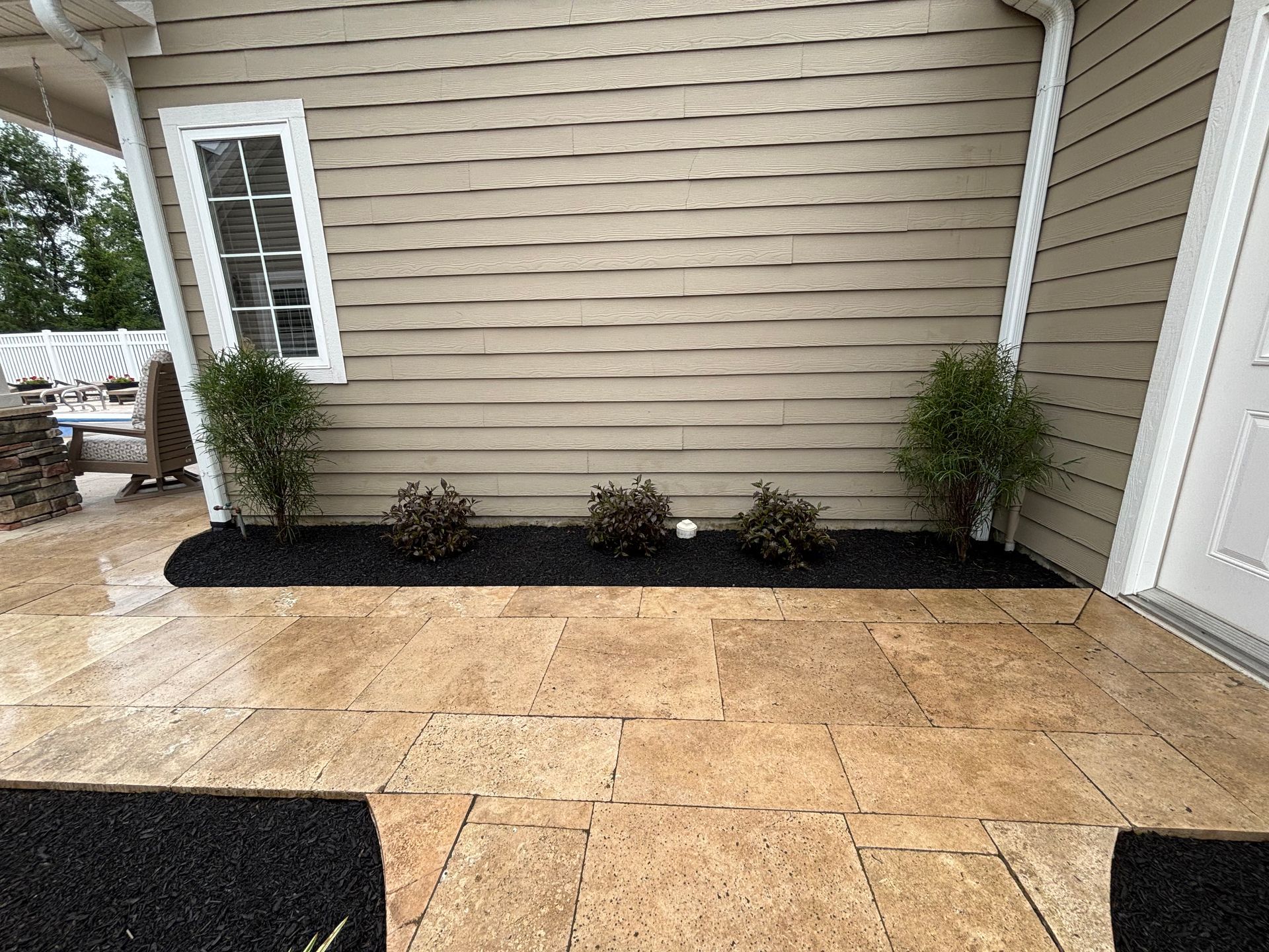 Patio with tan pavers, tan siding, small bushes, and a white door.