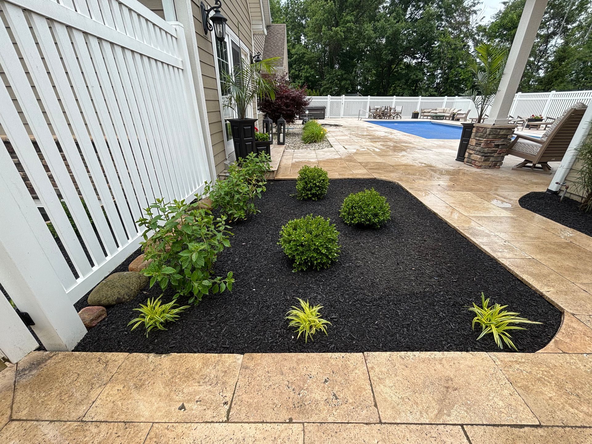 A black mulch flower bed with green bushes and a white fence next to a pool.