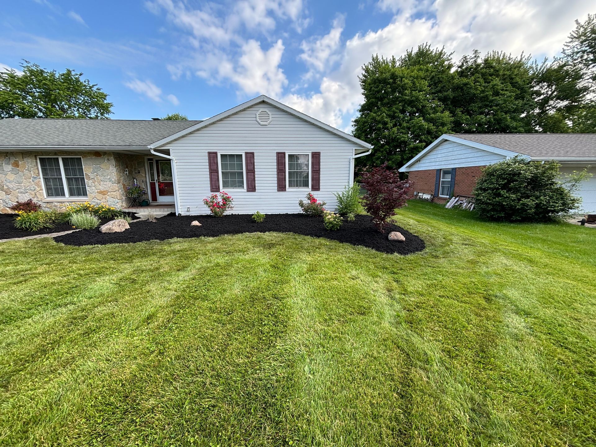 Small house with stone and white siding, dark mulch, red accents, and well-maintained green lawn under a blue sky.