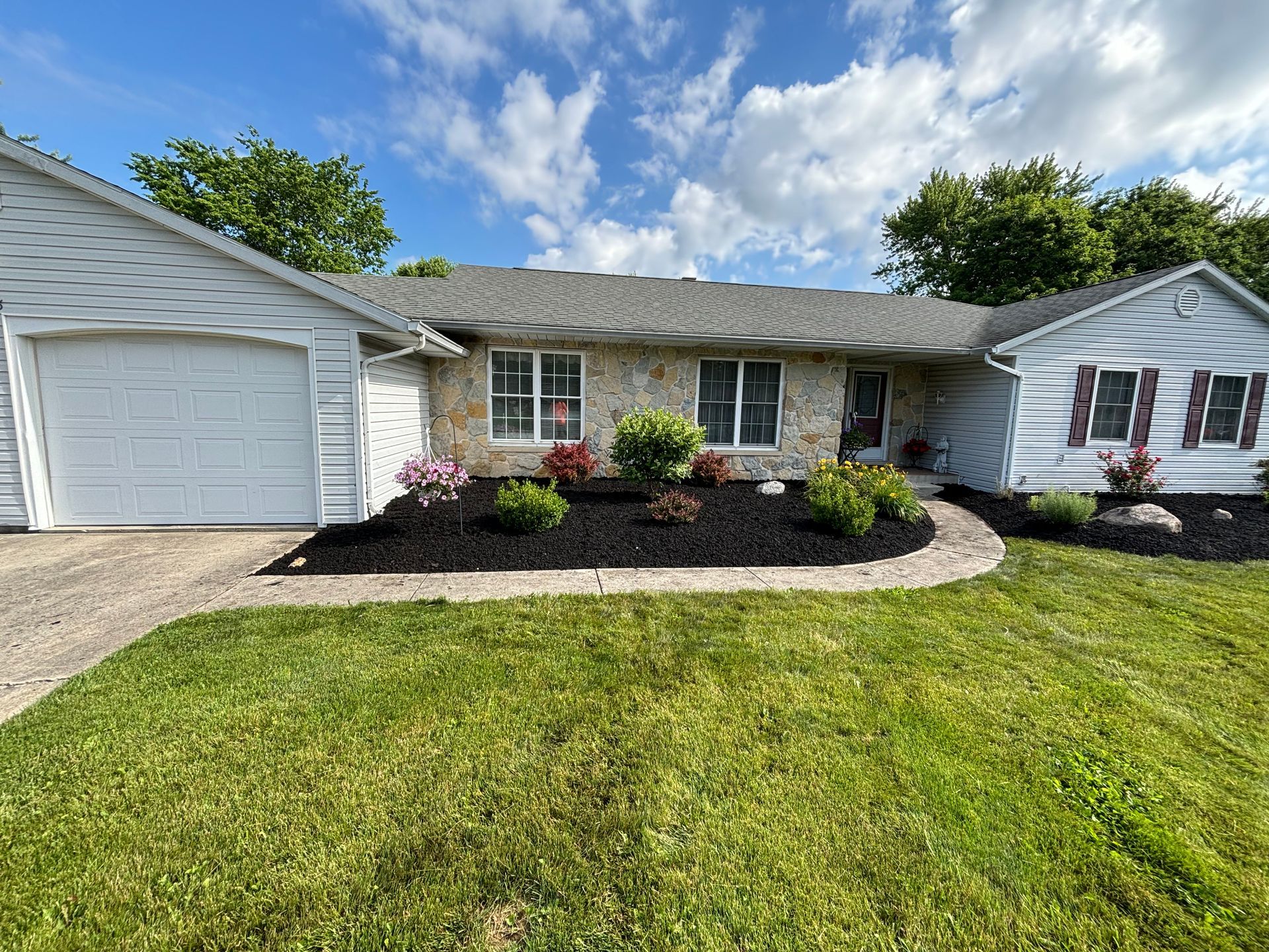 Ranch-style house with attached garage, dark mulch flowerbeds, and green lawn under a blue sky.