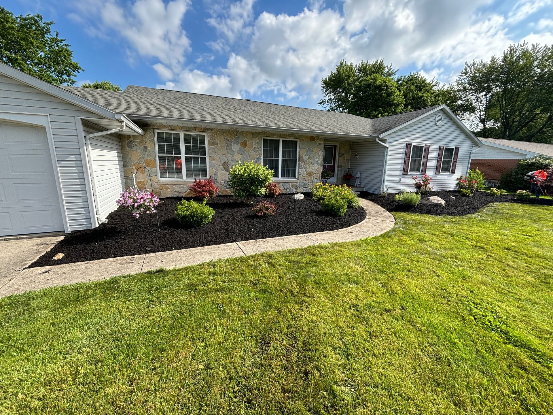 Ranch-style house with stone facade, black mulch flowerbeds, and green lawn under a partly cloudy sky.