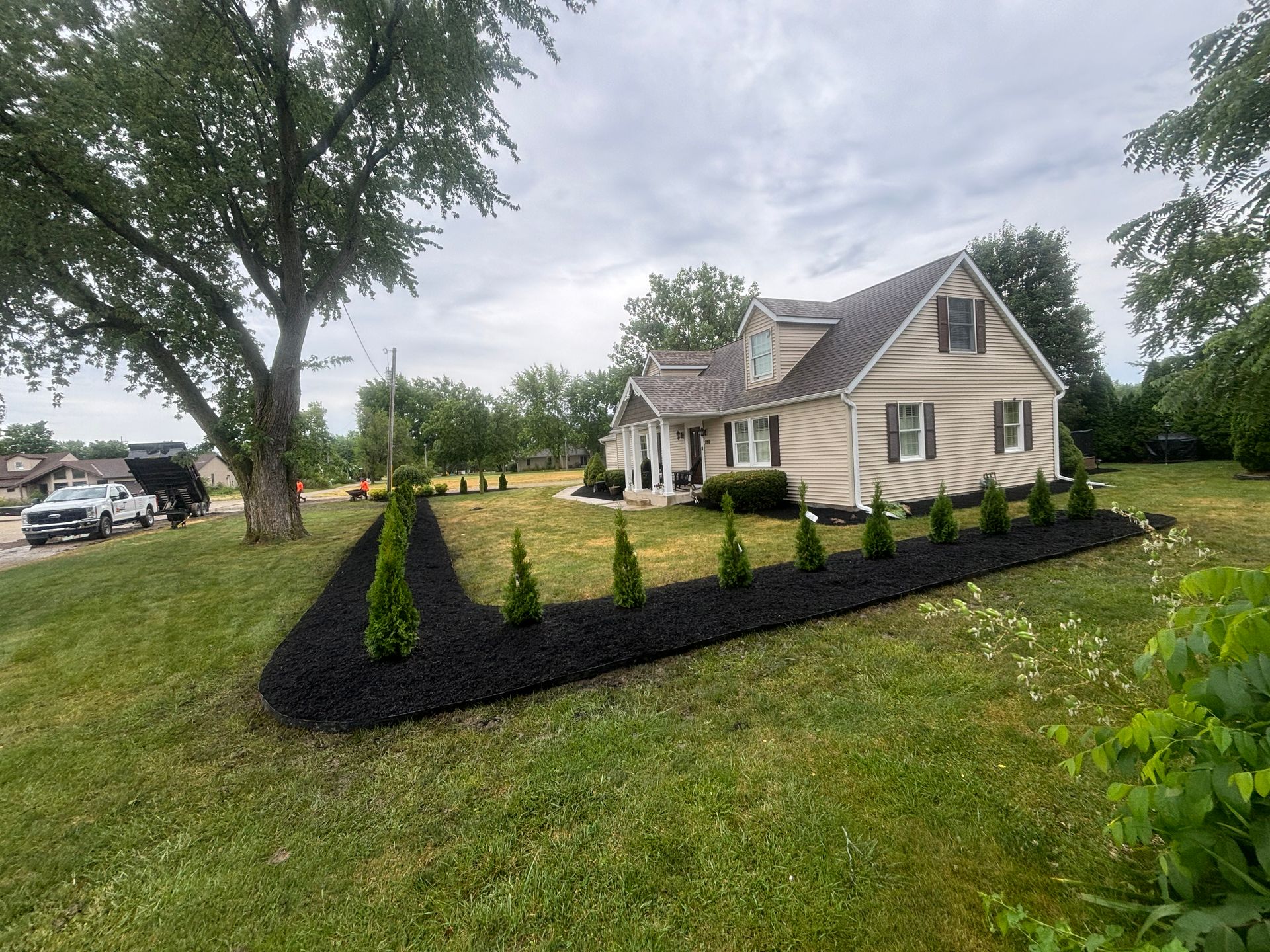 A tan house with black mulch beds and small green trees. A truck dumps mulch in the background.