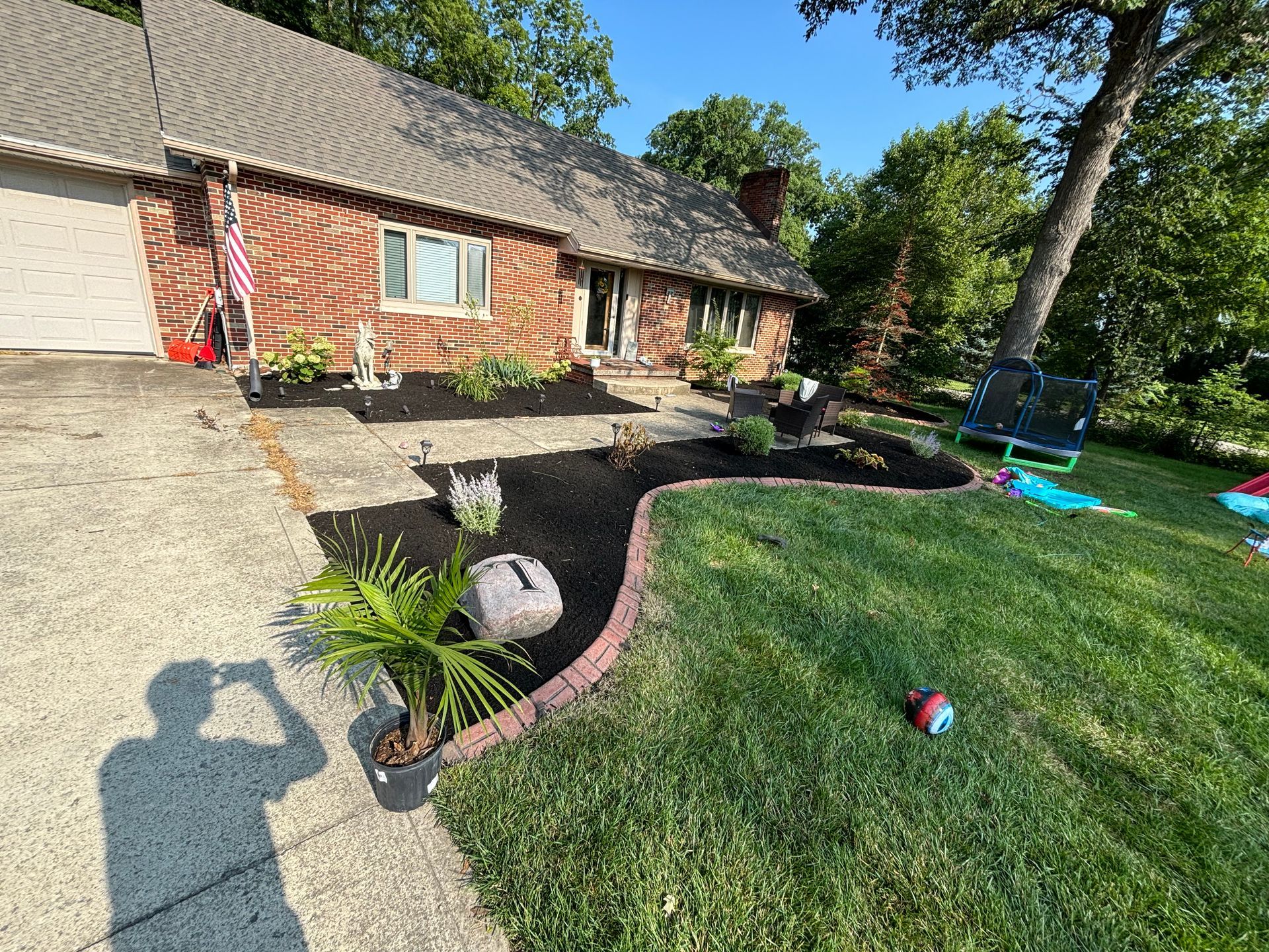 A house with a red brick facade, a driveway, and a landscaped garden. A person's shadow is in the foreground.