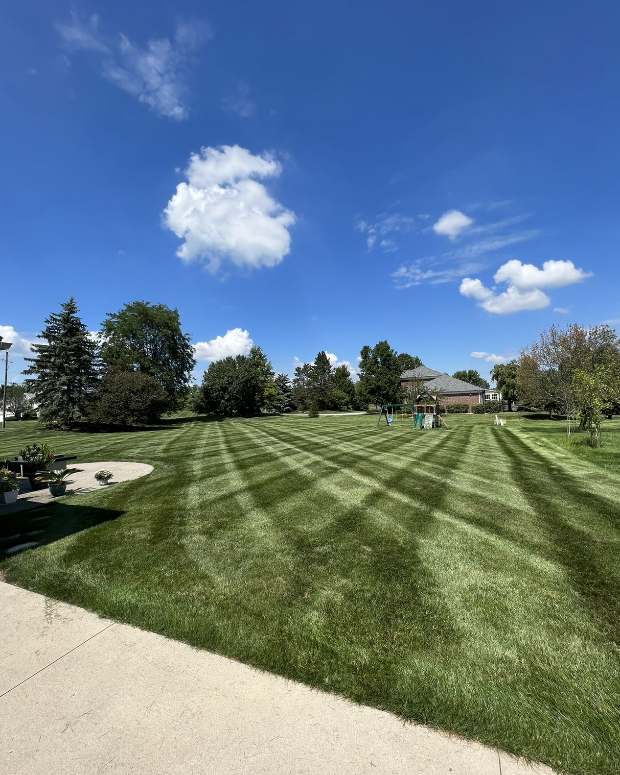 Lawn with geometric mowing pattern under a blue sky with puffy clouds, trees and a house in the distance.