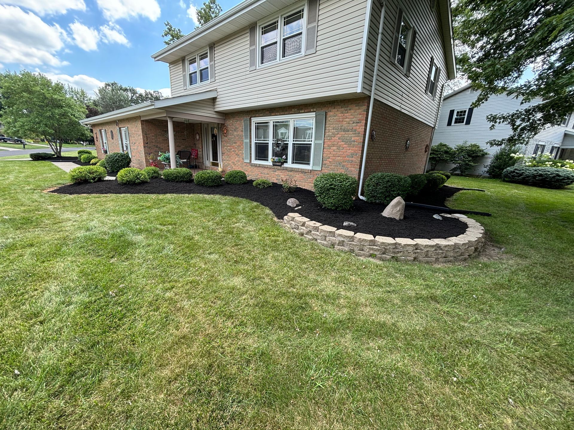 Two-story brick house with landscaped flowerbeds, green lawn, and stone retaining wall.