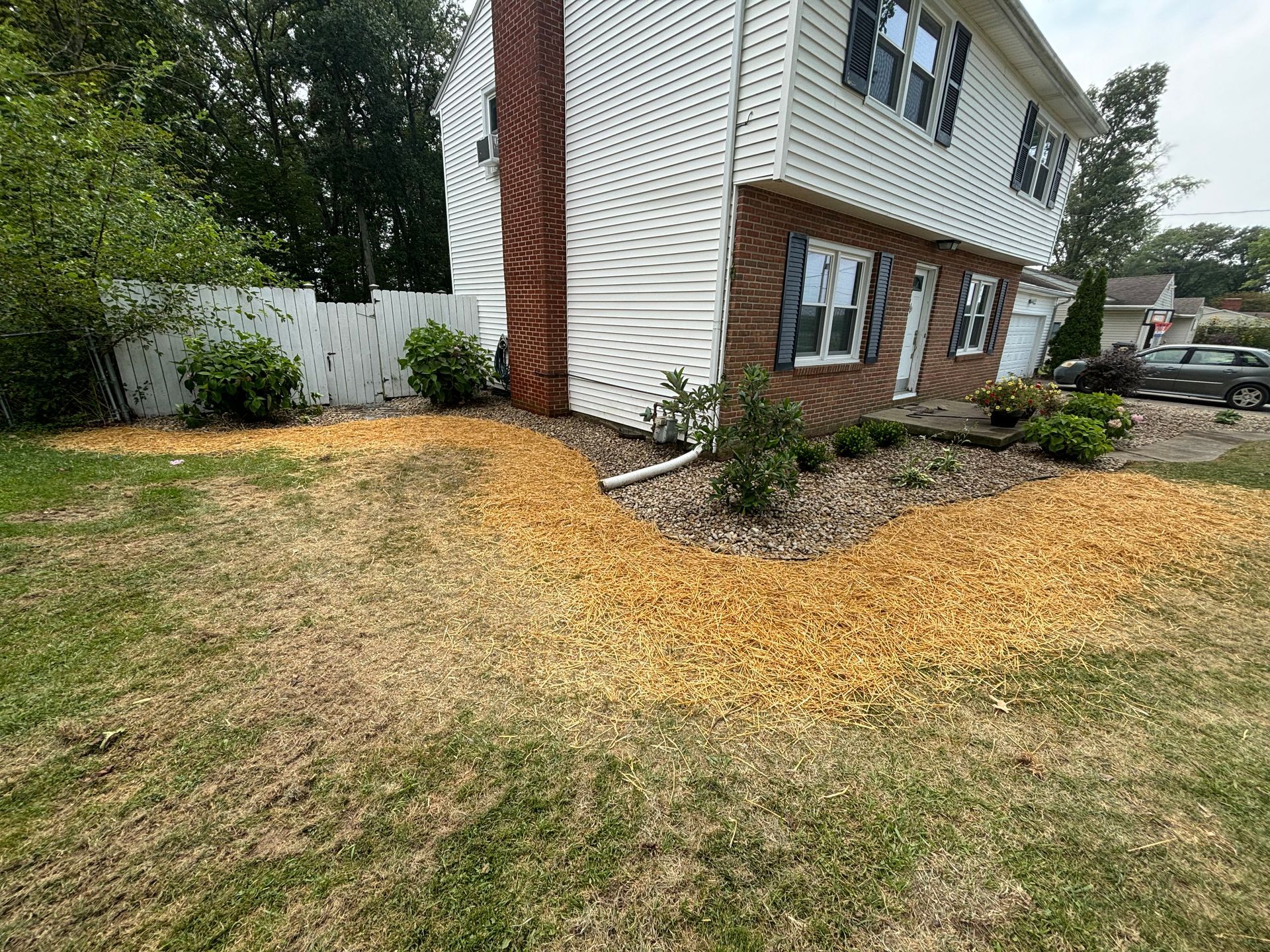 Two-story house with a brick chimney and landscaping. A path of wood chips leads to the front door, with some dead grass.