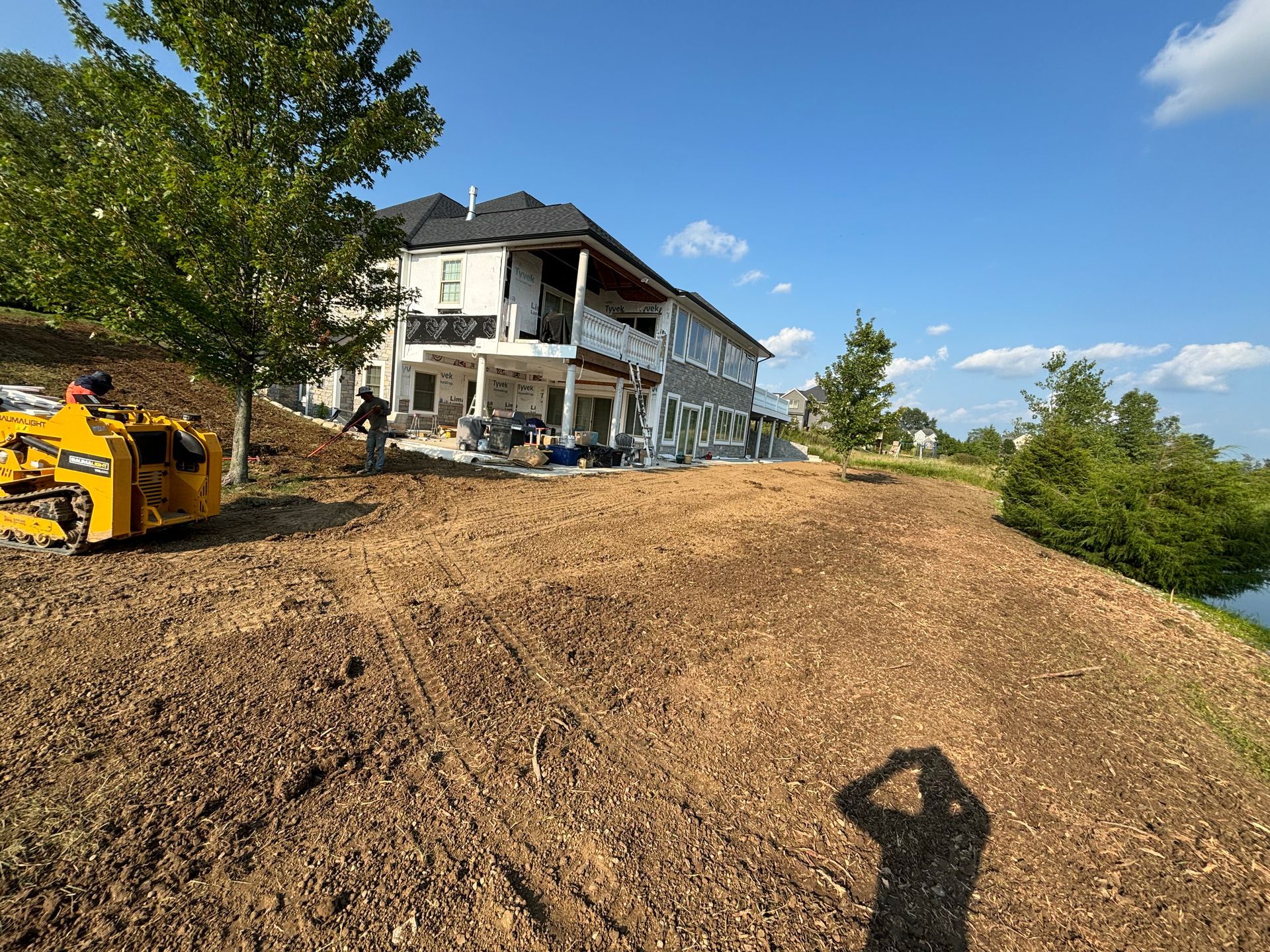A hillside being cleared with a yellow machine near a large house and small pond.