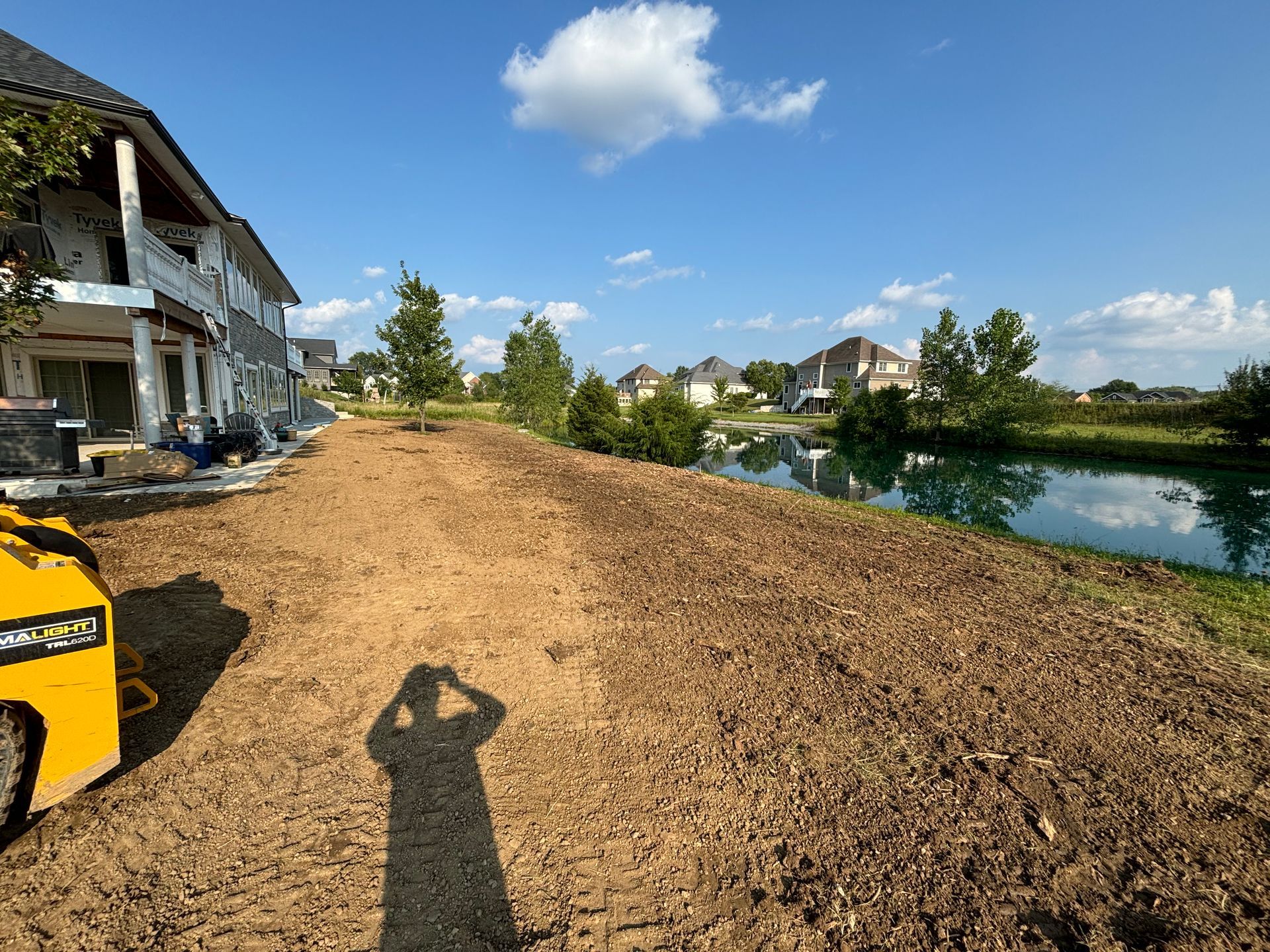 Construction site near a body of water with dirt and a machine, houses in the background, bright sky.