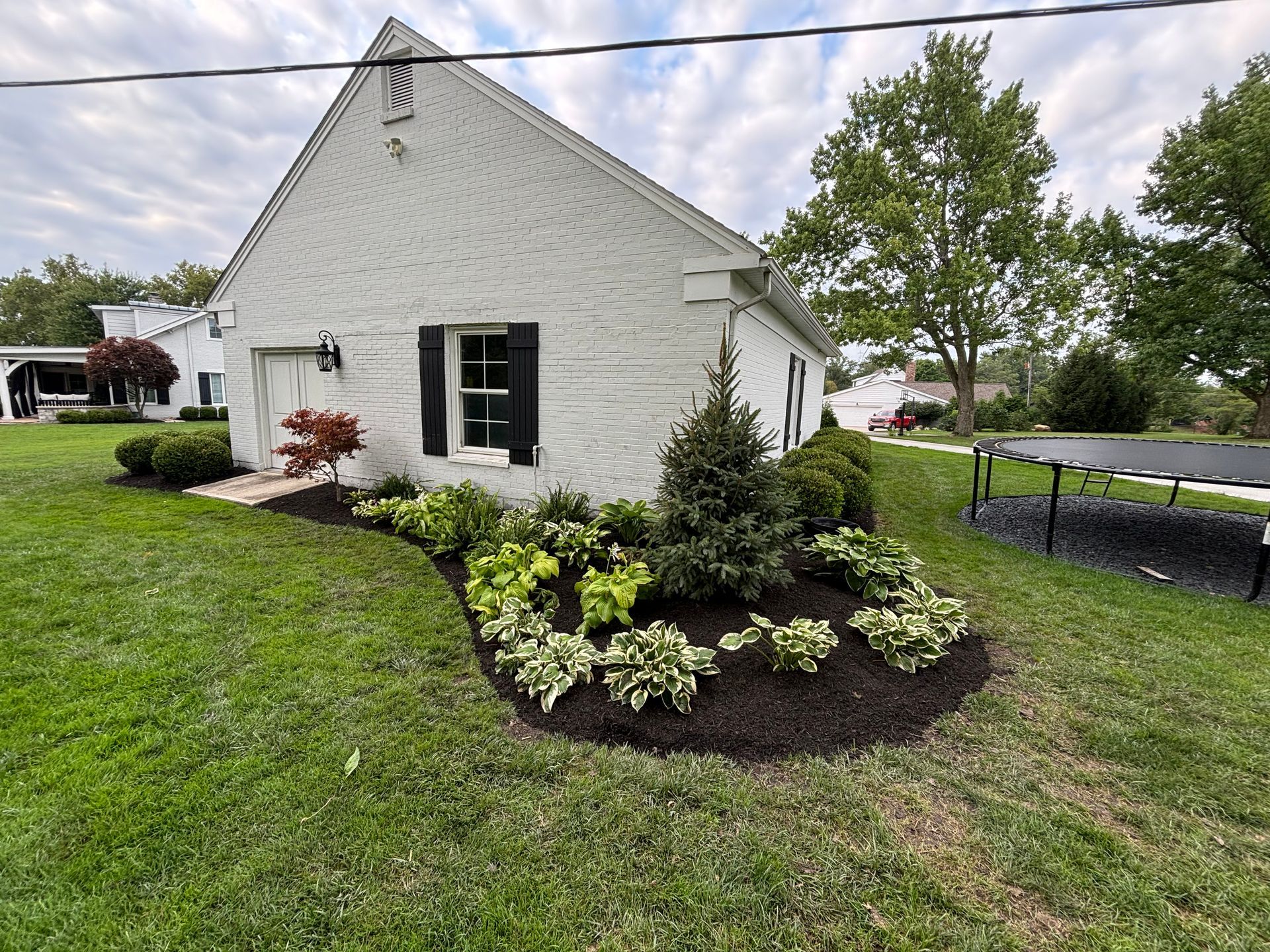 White brick house with landscaped garden and black mulch bed. Green grass lawn, cloudy sky.