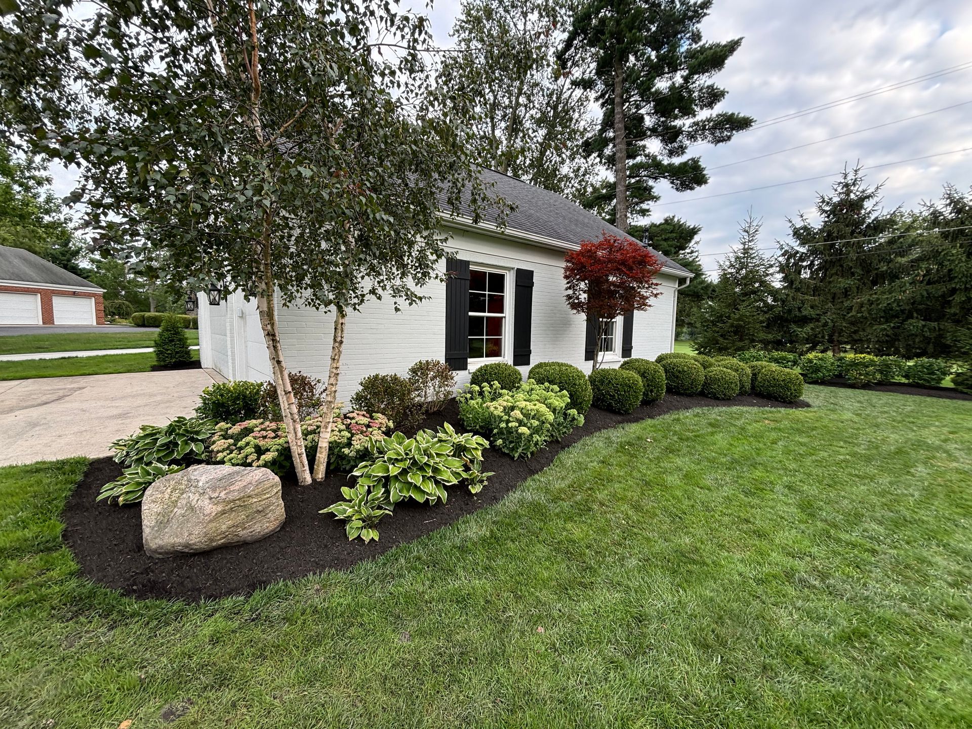 White building with black shutters, surrounded by lush green grass, trees, and a flower bed.