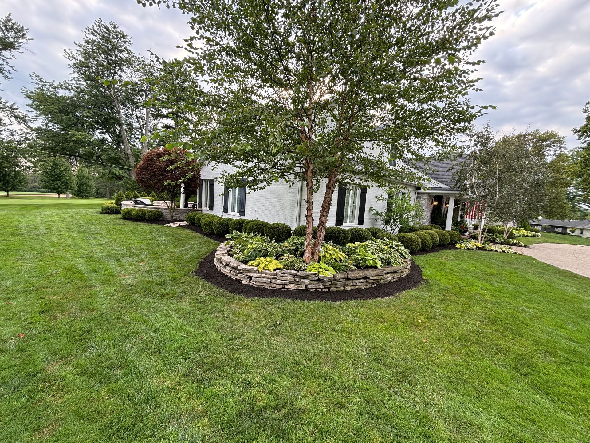 White house with landscaped yard, stone-bordered flower bed around a tree, green grass, and cloudy sky.