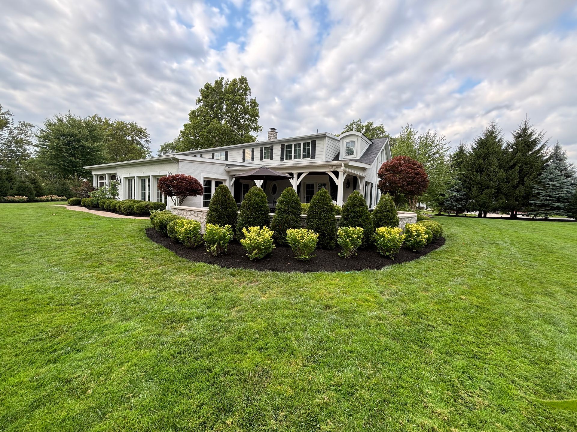 A white house with a wraparound porch, surrounded by green grass and a curved shrub bed. Overcast sky.