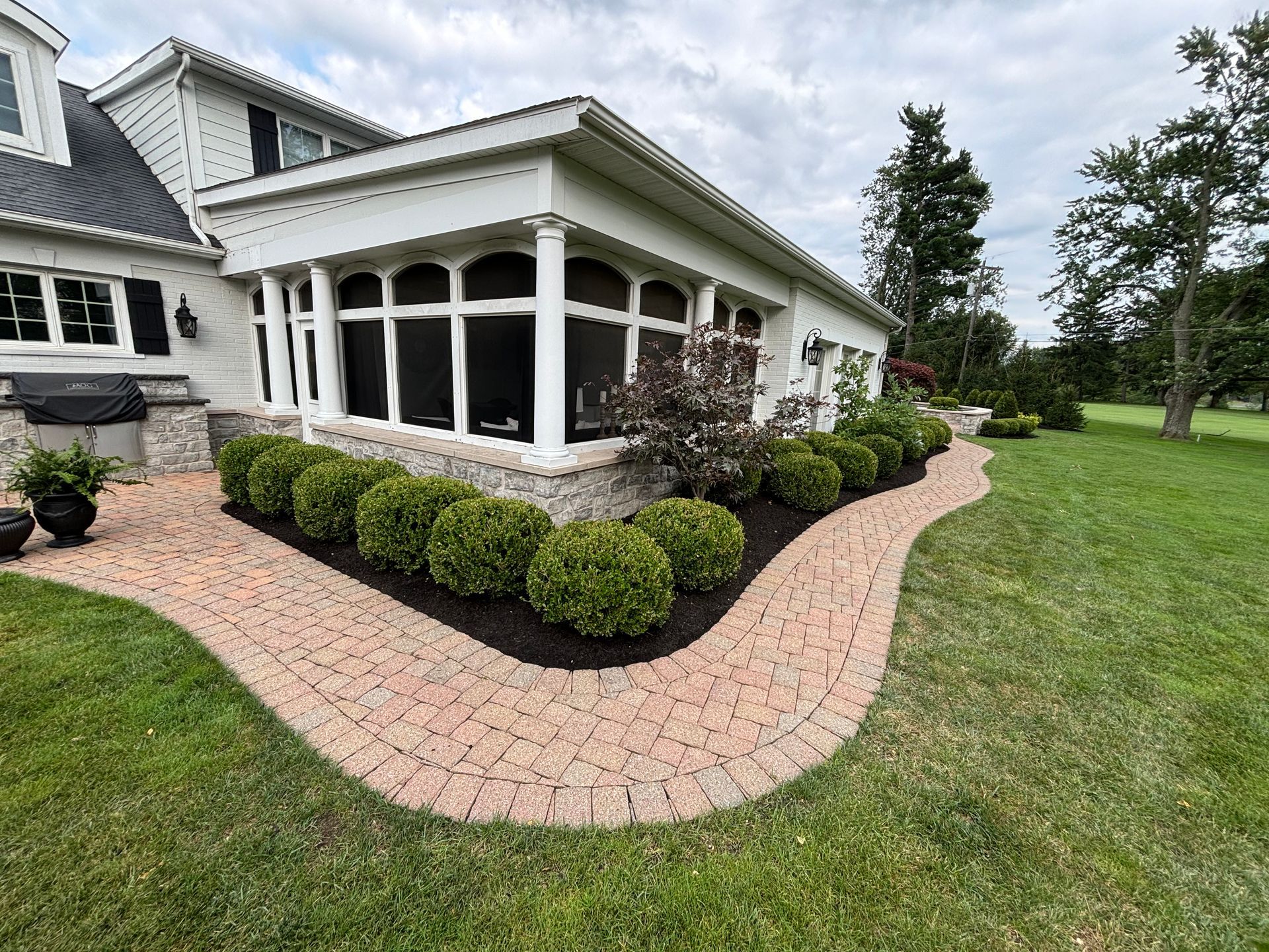 Brick pathway curves around a white patio with a hedge of round green bushes, on a grassy lawn.