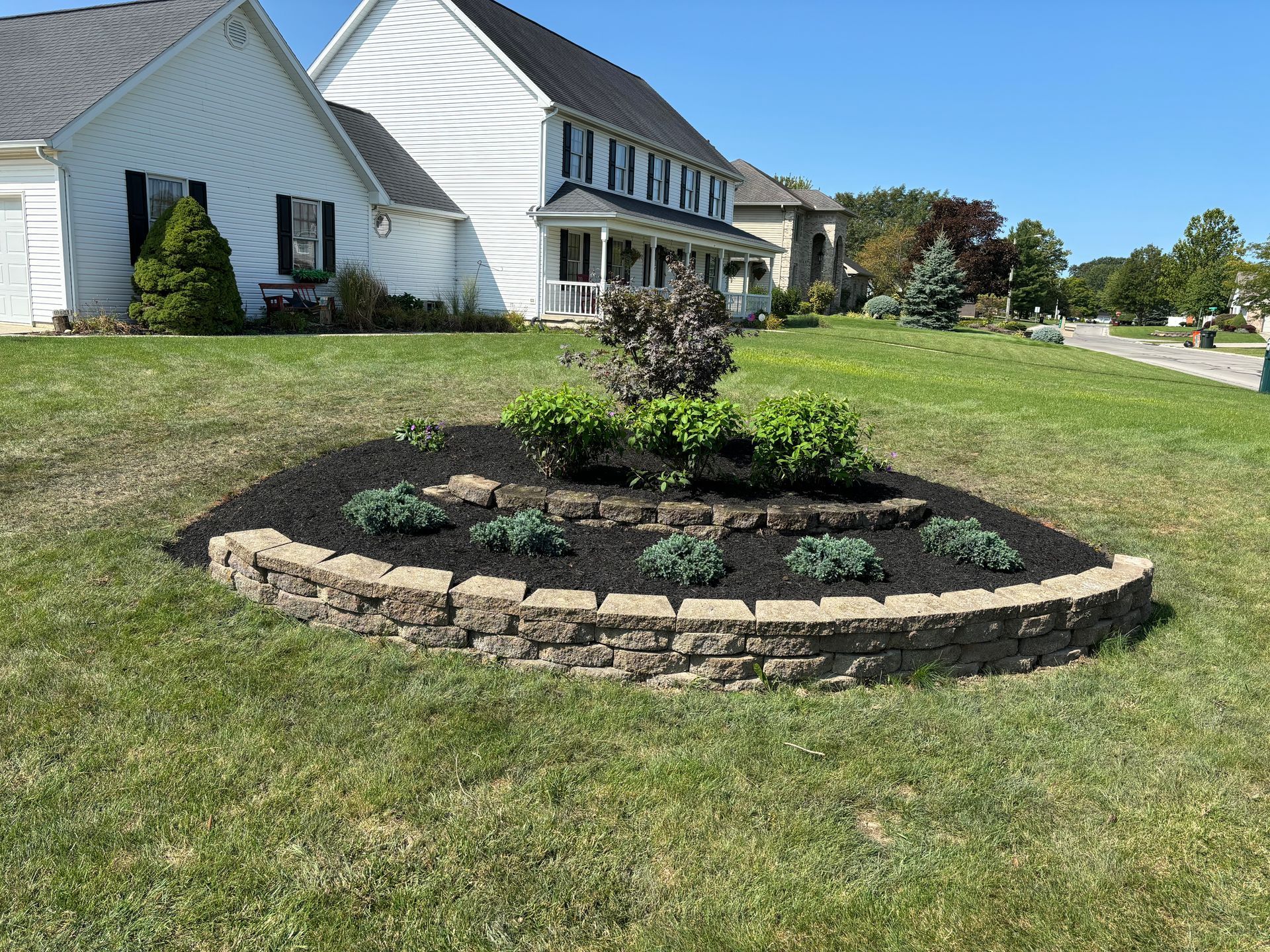Raised flower bed in front yard with brick edging, mulch, and various plants.