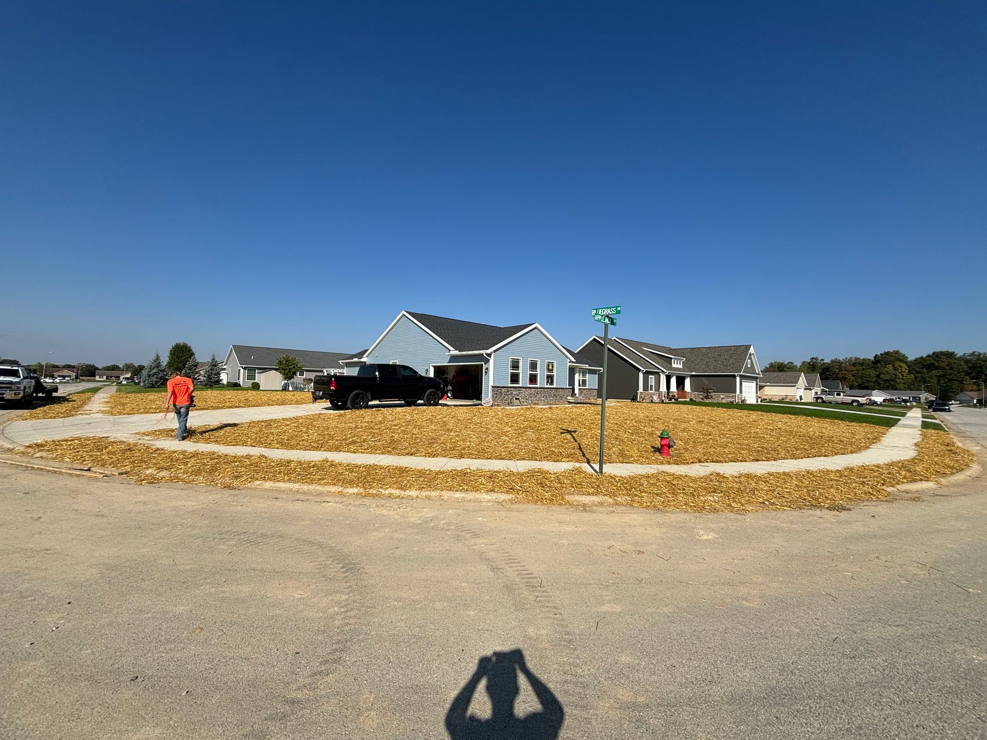 A person on a street applies mulch to a large, circular landscape bed in front of houses under a blue sky.