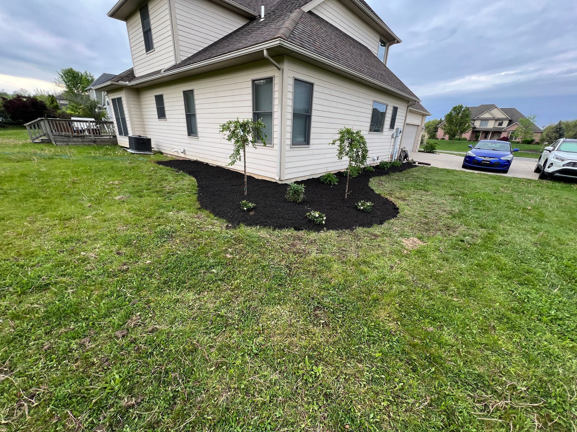 House with dark mulch landscaping, small trees, and cars in the driveway. Green grass in front.
