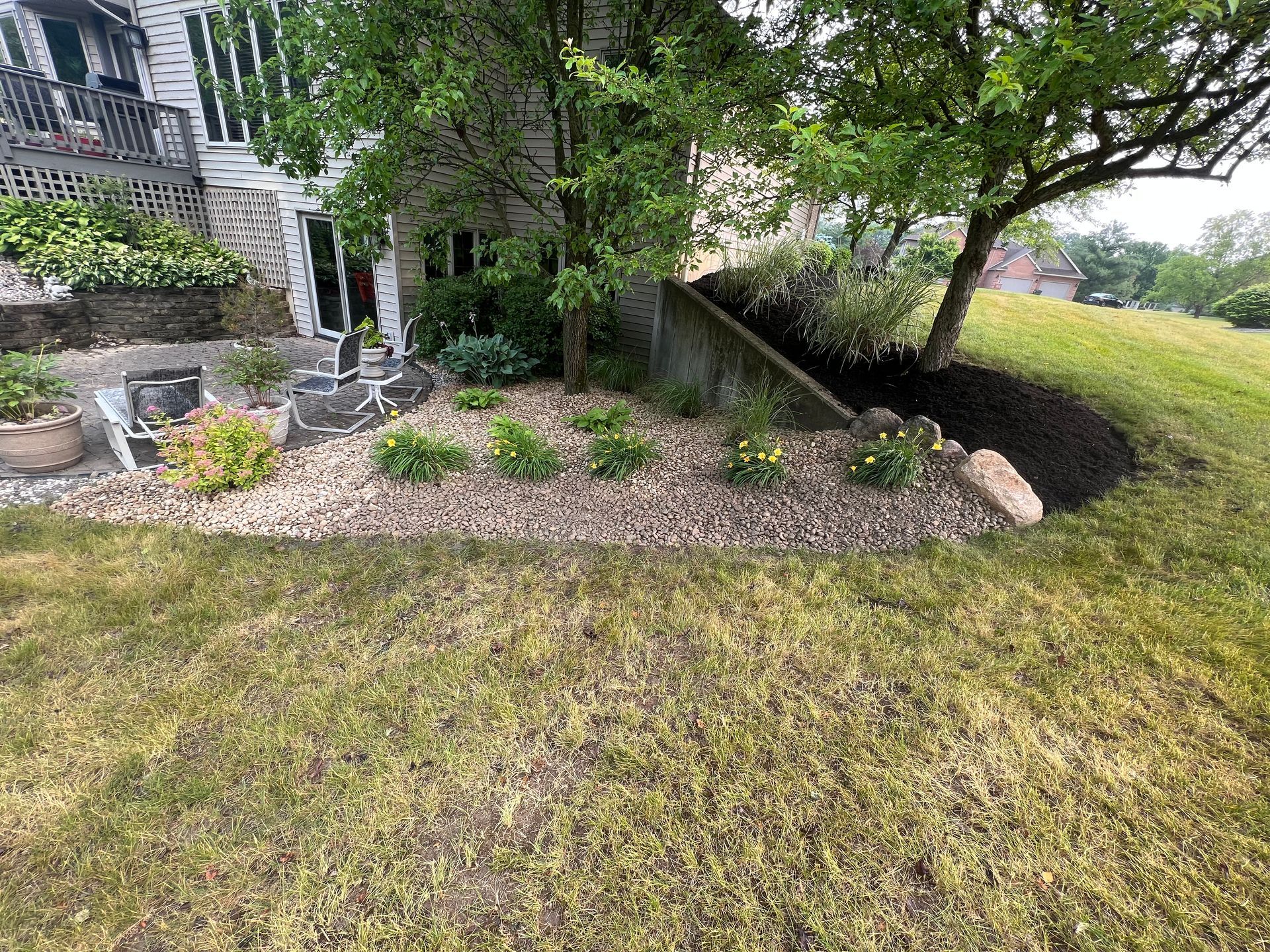 Lawn with a garden bed of rocks and plants next to a house, and a tree on a hill.