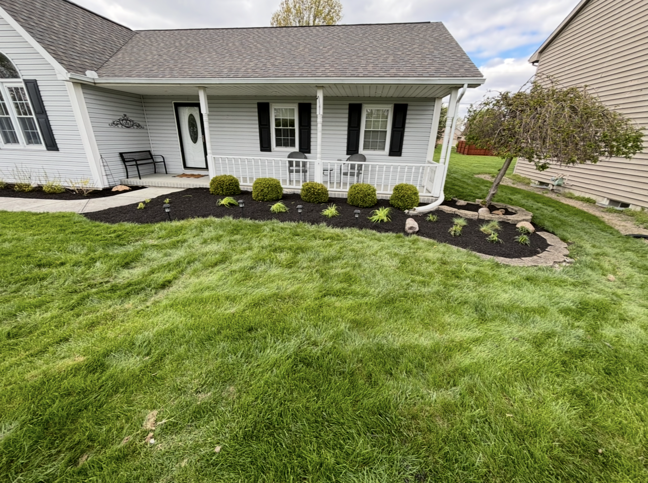 A house with a gray roof, white porch, and a green lawn, a flower bed with bushes, and a small tree.