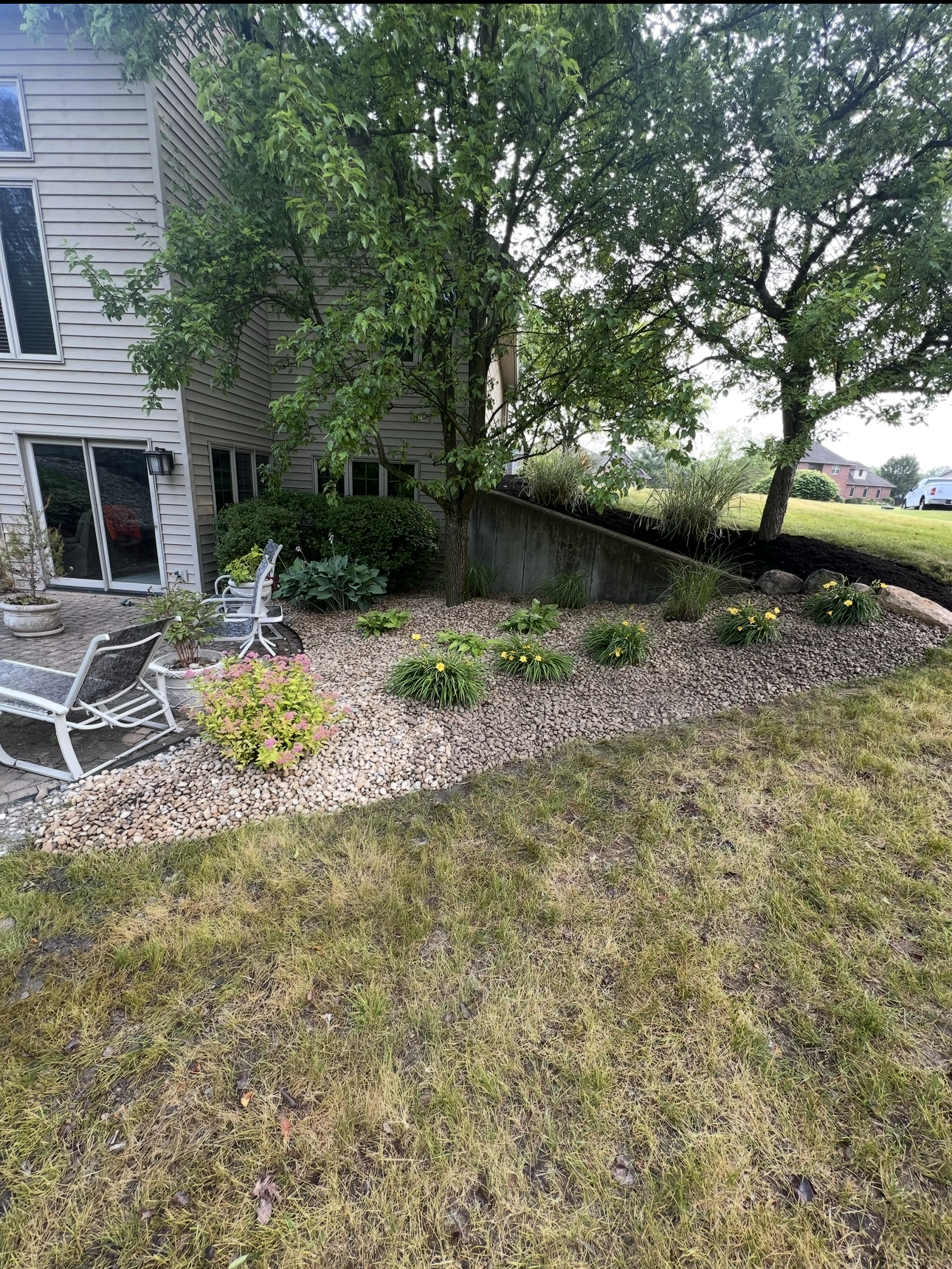 Exterior view of a house with a yard, flowerbeds, and two chairs. The lawn is dry, and the sky is overcast.