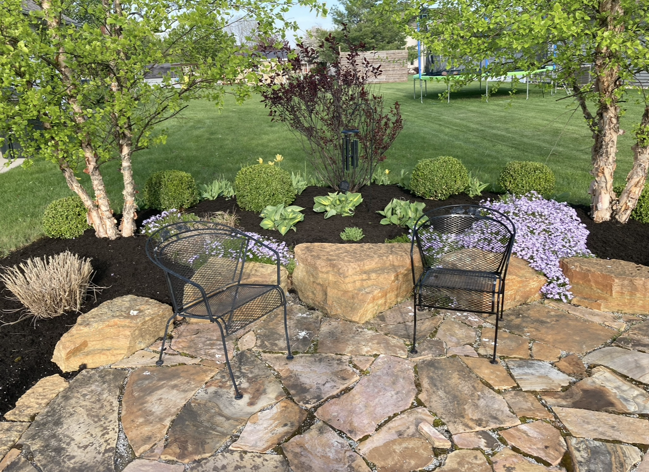 Stone patio with black chairs, boulders, and a landscaped garden with trees.