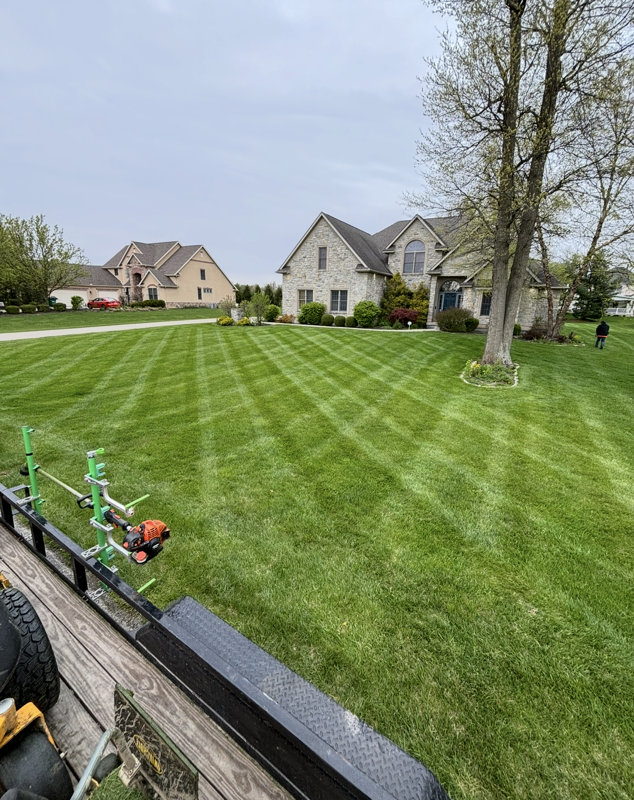 Lawn mowed with stripes in front of a stone house, trees, and a cloudy sky. Lawn care equipment in foreground.