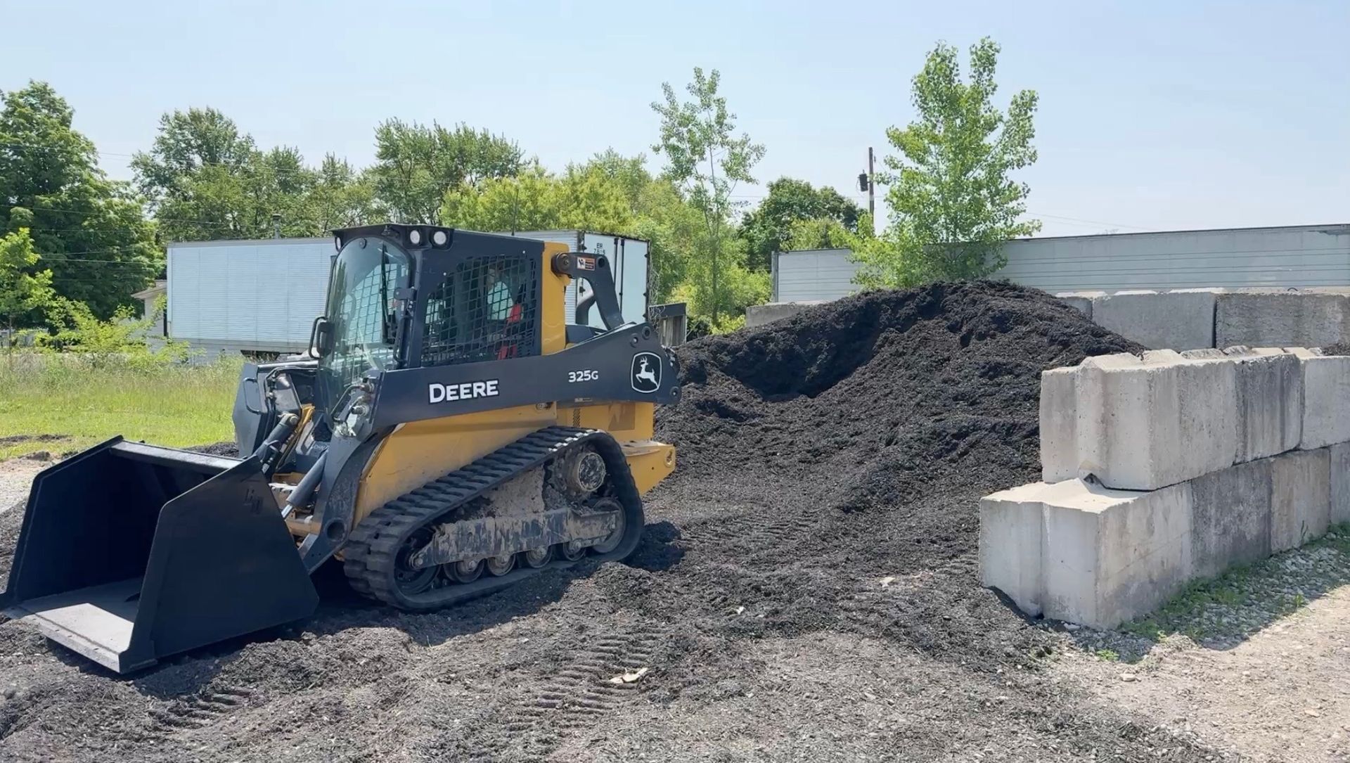 A yellow and black skid steer loader scooping up dark mulch in a construction area on a sunny day.