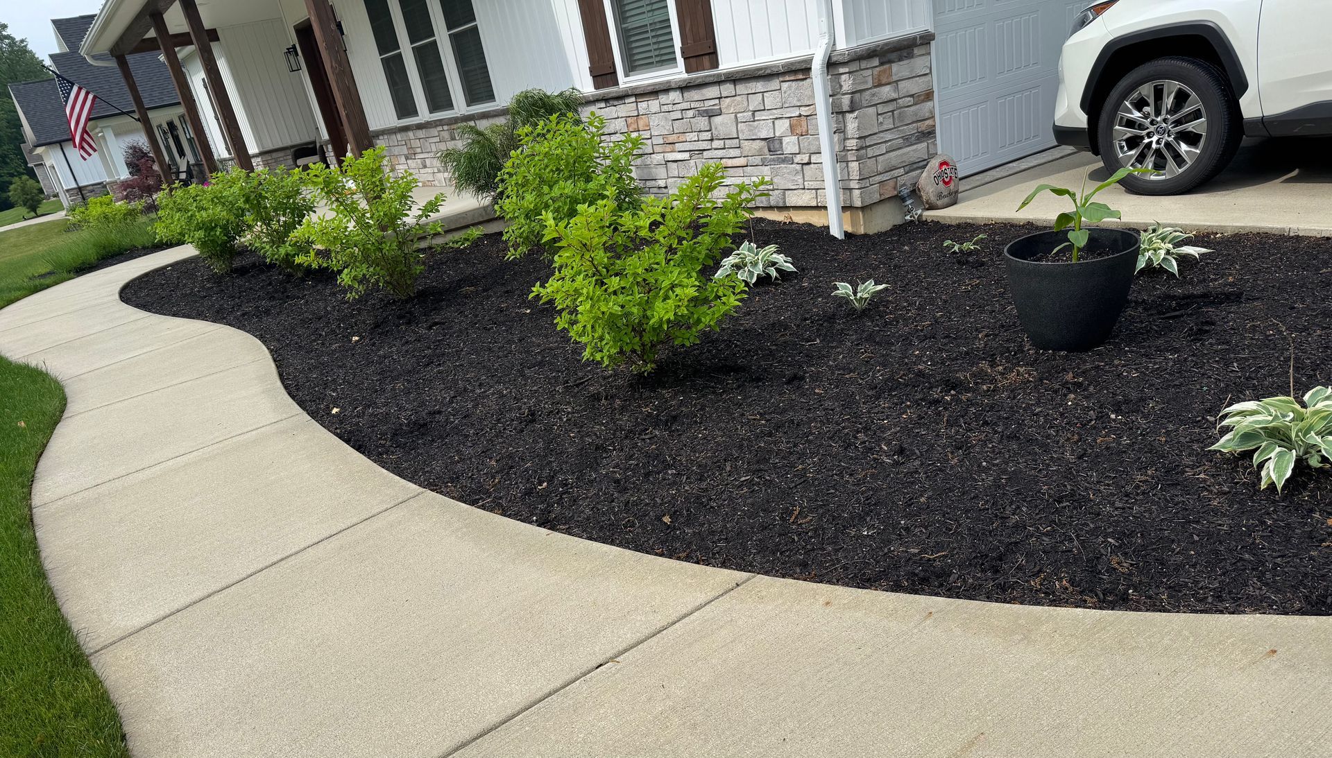 Curving sidewalk leads to a house with dark mulch flower beds, green bushes, and a black pot.