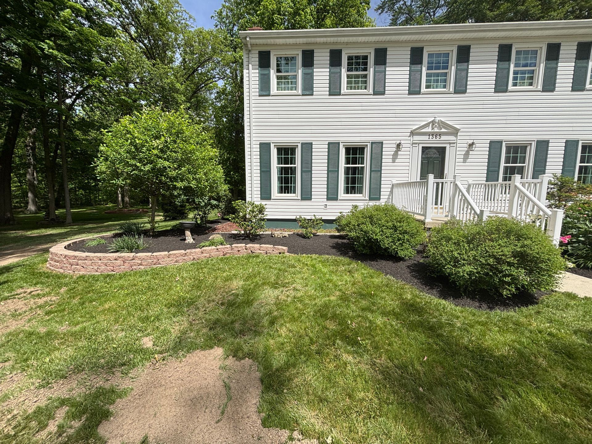 White house with green shutters, front yard with flowerbeds, grass, and trees.