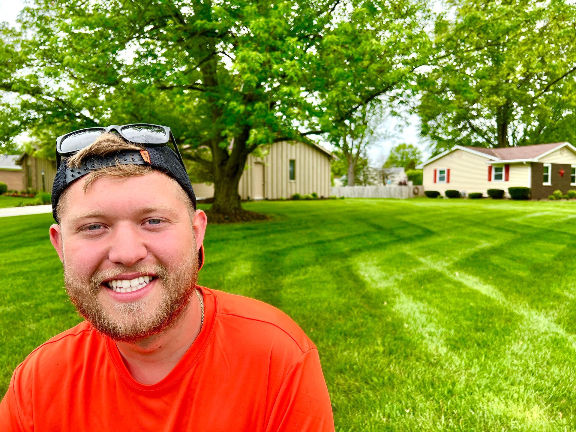 Man smiling in front of freshly mowed lawn, wearing a hat and orange shirt. Green grass, trees, and houses.