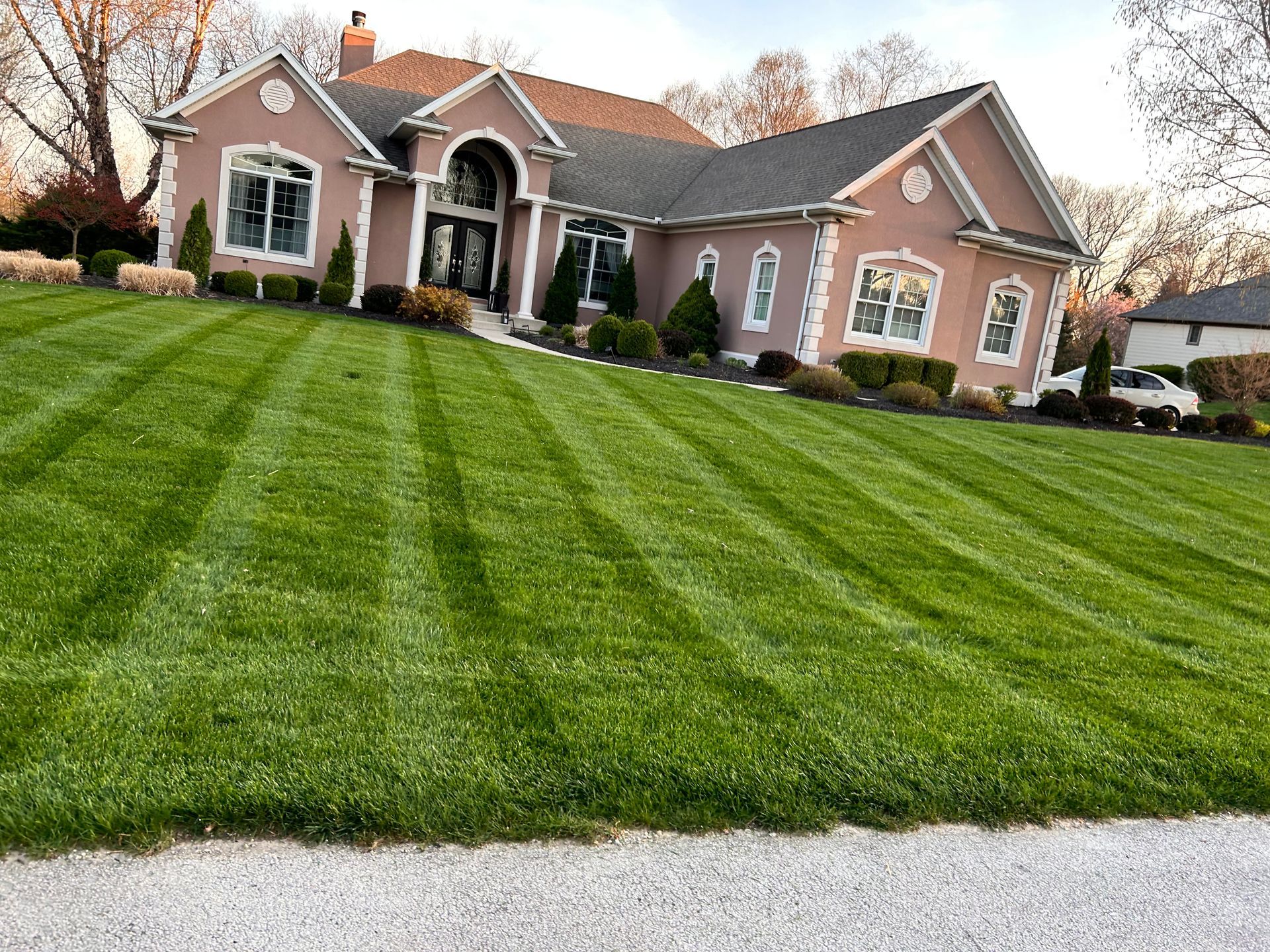 Lush green lawn striped with mower lines slopes up to a large pink house with a manicured landscape.