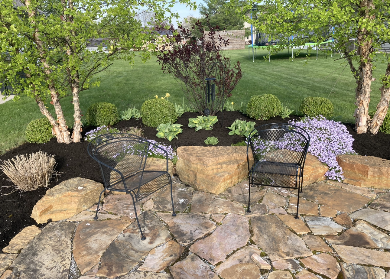 Patio with two black chairs, large stones, garden bed, and trees.