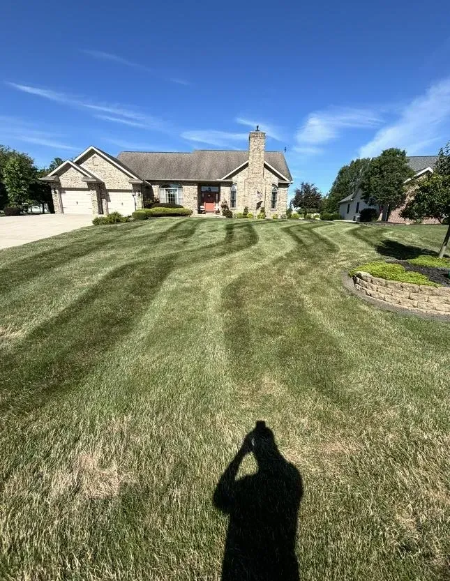 Lawn mowed in striped pattern, with trees and a gravel driveway in the background. Sunlight casts shadows on the grass.