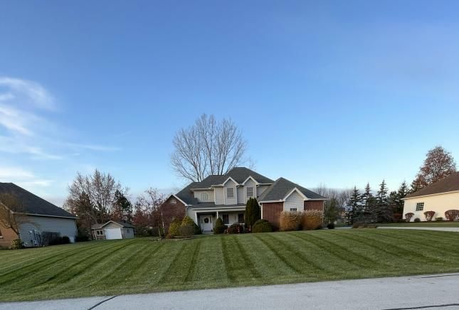 Green lawn with geometric mowing patterns, a pond in the background, and a sunny sky. Logo in the bottom right corner.