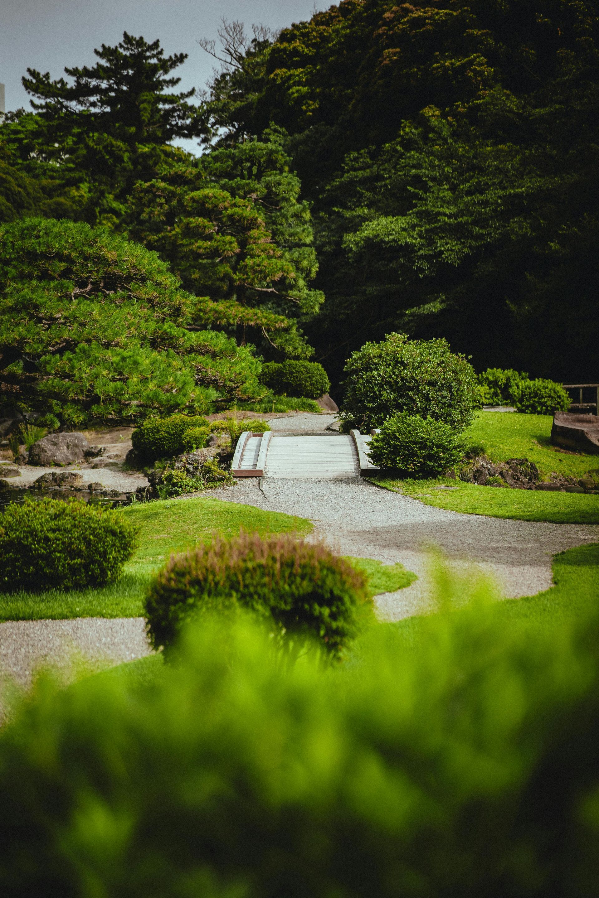 Lush green garden with a gravel path, trimmed shrubs, and a small white bridge in the center
