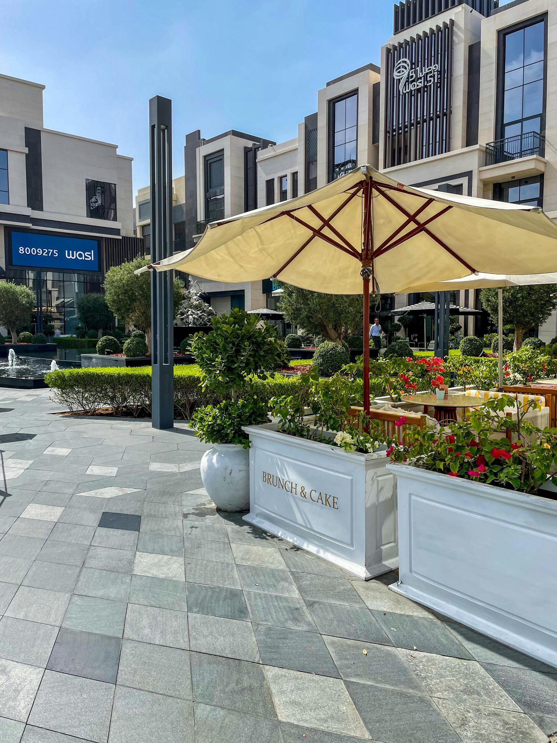 Sunny outdoor café patio with beige umbrellas, greenery, and modern buildings in the background