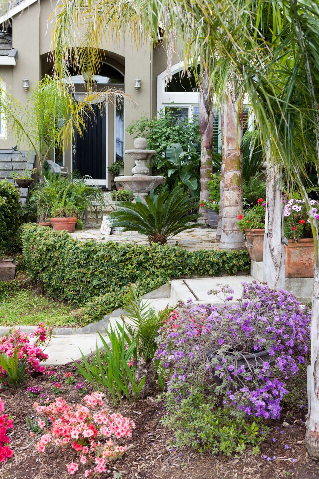 A well-landscaped front yard with a fountain, colorful flowers, and palm trees.