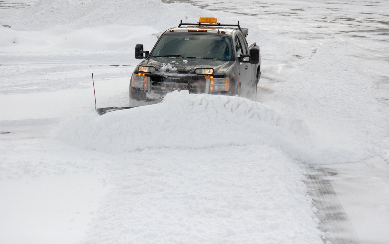 Snowplow truck clearing a snow-covered road. The truck is dark-colored, with a yellow light on top, pushing a large amount of fresh snow.