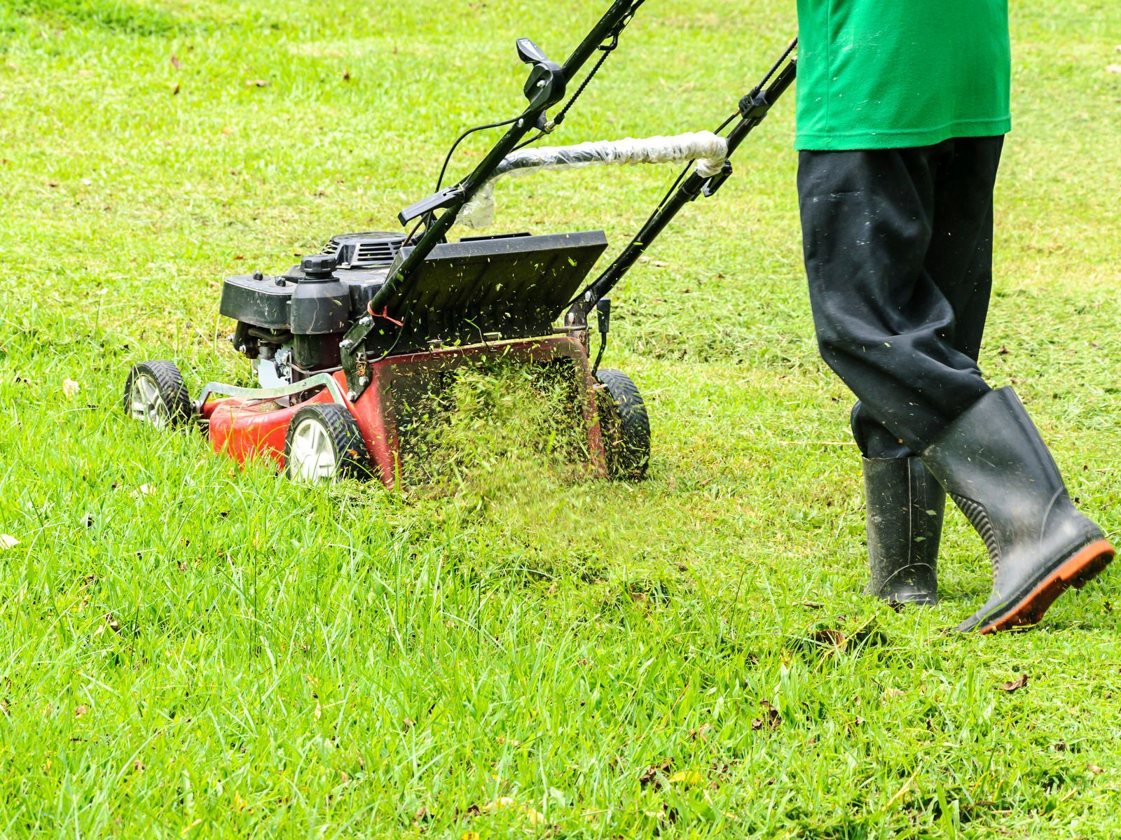 Person mowing green grass with a red lawnmower, wearing a green shirt and black boots.