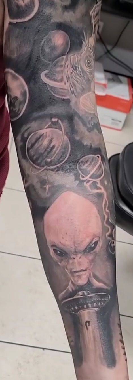 Tattoo of an alien on a spaceship with planets and galaxies on a forearm.