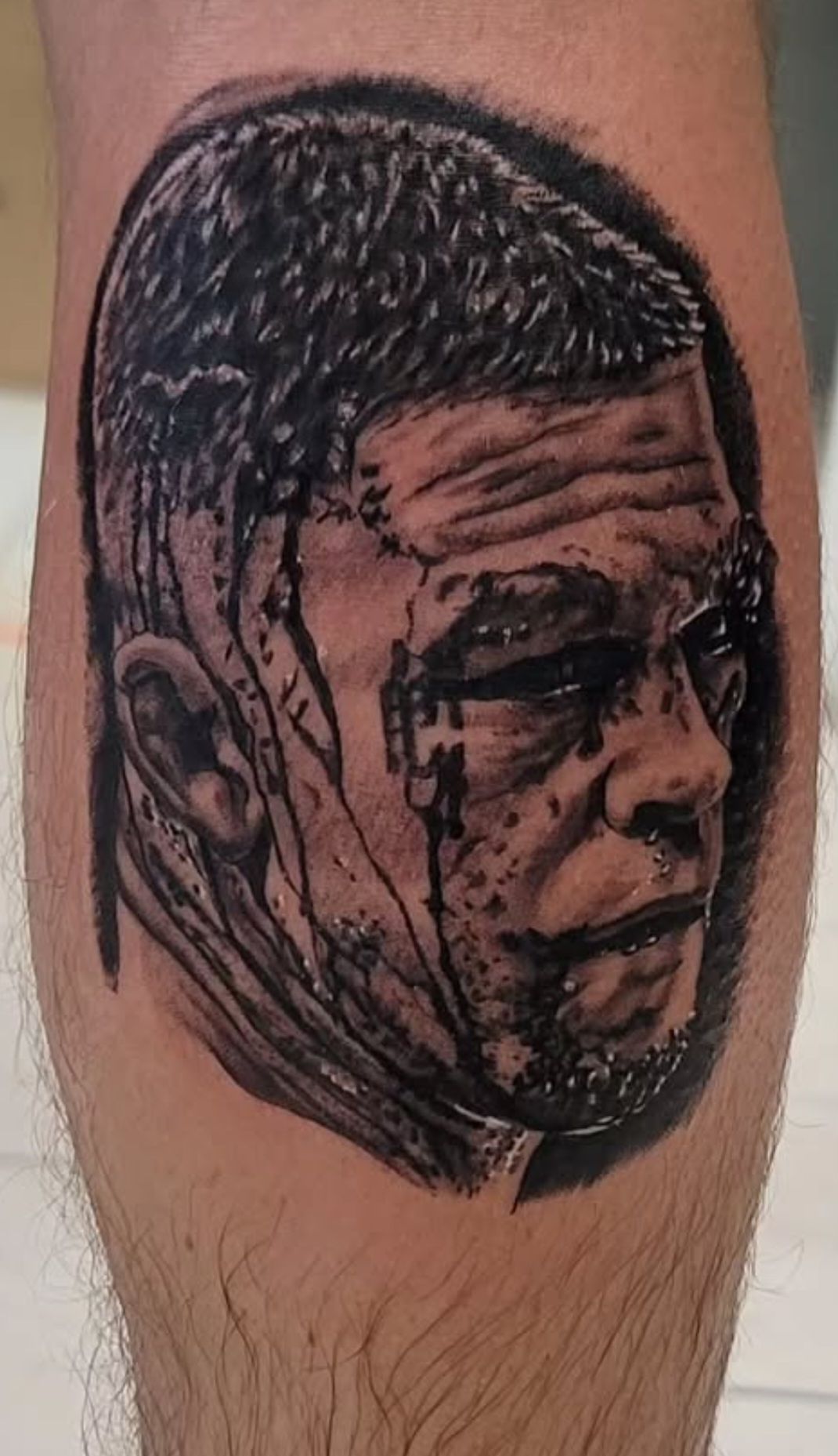 Tattoo of a man's face, with a bloody, bruised appearance. Black and gray ink on a leg.