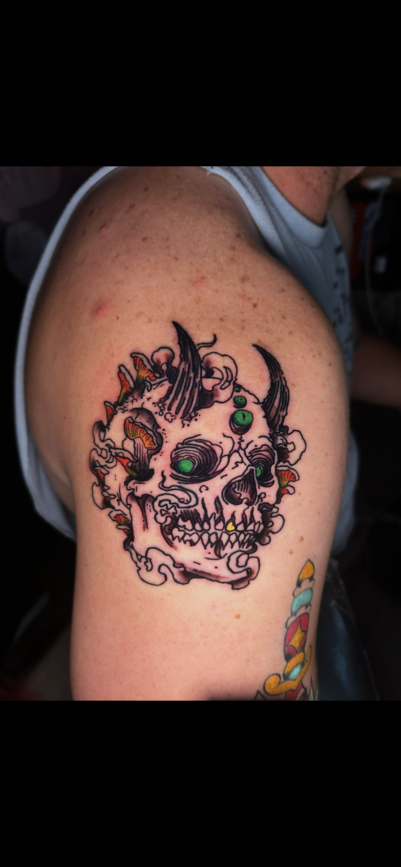 Tattoo on shoulder, a stylized skull with green eyes and horns, surrounded by floral elements.