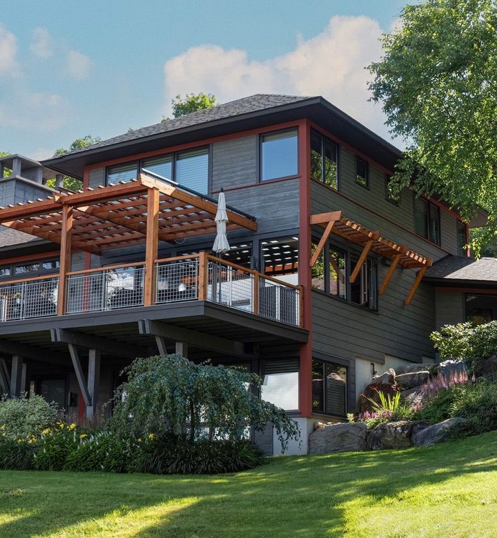Modern two-story house with wood deck, pergola, and lush green lawn on a sunny hillside