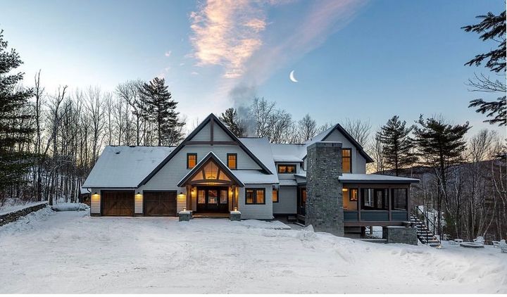 Snowy modern chalet at dusk with lit windows, stone chimney, and crescent moon over bare trees