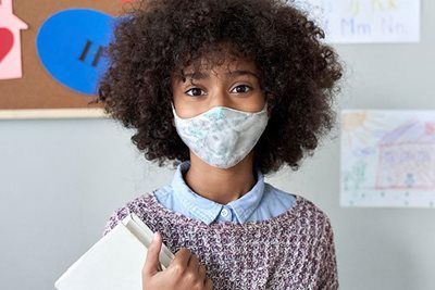 Young person with curly hair, wearing a mask and holding a book in a classroom setting.