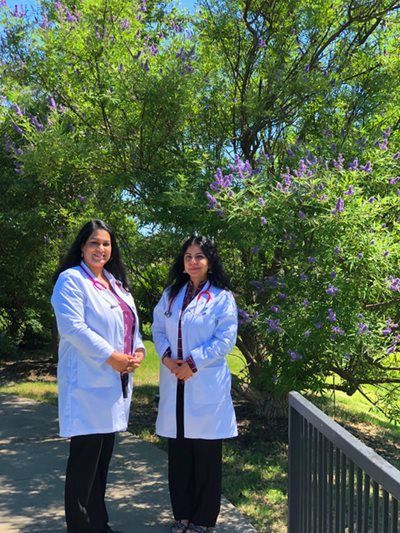 Two women in white lab coats stand outside, smiling. Purple flowers bloom in the background.