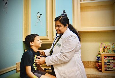 Doctor listening to a child's chest with a stethoscope in an exam room. The child smiles.